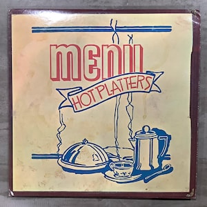 May include: A vintage menu sign with a cream-colored background and a dark brown border. The word "MENU" is in red block letters, and "HOT PLATTERS" is in blue on a banner. Blue illustrations of a covered dish, coffee pot, and cup.