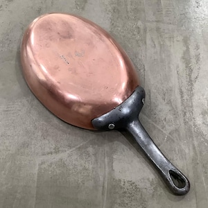 May include: A copper oval-shaped pan with a long, dark metal handle. The pan's interior is a shiny, reflective copper color, while the handle and the area where it connects to the pan are a darker, matte metal. The pan appears to be vintage or antique.