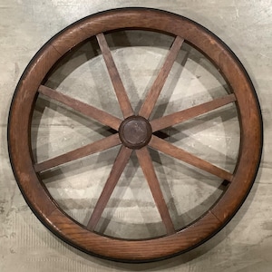 May include: A wooden wagon wheel with a dark brown rim and spokes. The wheel has a central hub and is made of wood. The spokes radiate outwards from the center, creating a classic, rustic design. The wheel is likely an antique or vintage item.
