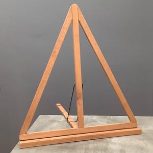 May include: A wooden easel with a triangular A-frame design. The easel is made of light-colored wood and features a black metal support. The base is rectangular, and the easel is designed to hold artwork or display items.