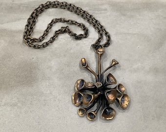 May include: A bronze necklace with a pendant shaped like a stylized flower with multiple petals. The chain is made of linked circles.