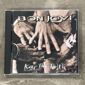May include: A Bon Jovi "Keep The Faith" CD album in a clear jewel case. The album cover features a sepia-toned image of hands clasped together. The band's name is at the top, and the album title is at the bottom.