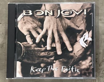 Op de afbeelding: Een Bon Jovi "Keep The Faith" cd-album in een doorzichtige jewelcase. De albumhoes toont een sepia-afbeelding van handen die elkaar aanraken. De bandnaam staat bovenaan en de albumtitel onderaan.