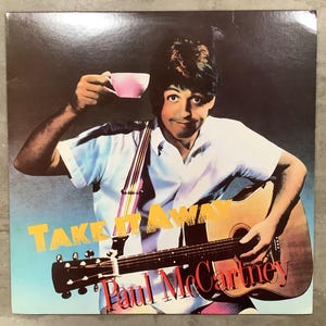 May include: Album cover featuring Paul McCartney holding a guitar and a pink teacup. The album title "Take It Away" is in yellow and the artist's name, Paul McCartney, is in red. The background has a gradient of blue and black.