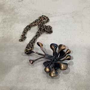 May include: A gold-toned necklace with a pendant shaped like a stylized flower with multiple petals. The pendant is made of a dark metal and has a textured surface.