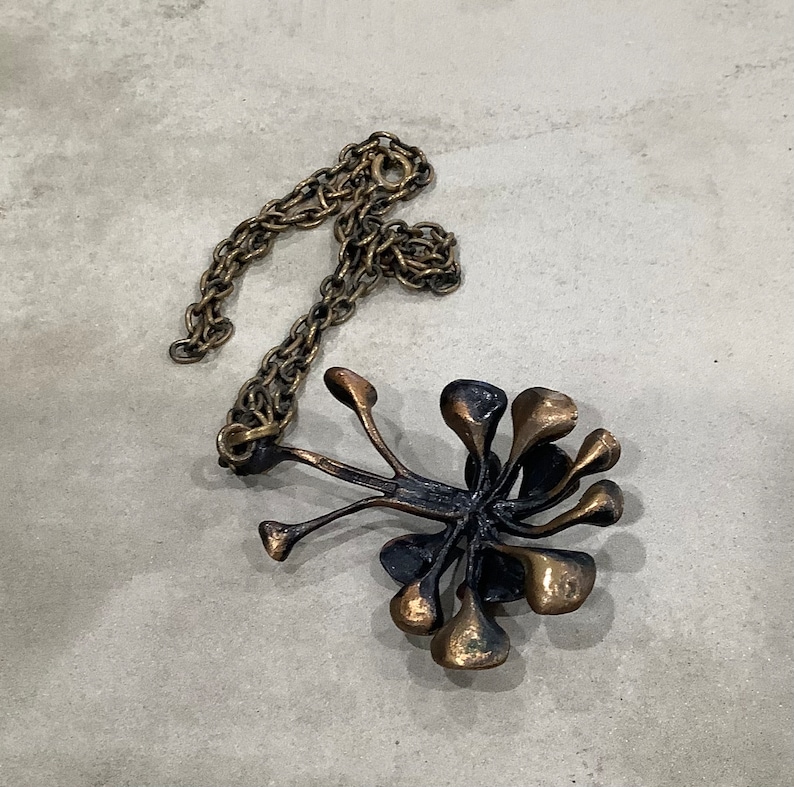 May include: A gold-toned necklace with a pendant shaped like a stylized flower with multiple petals. The pendant is made of a dark metal and has a textured surface.
