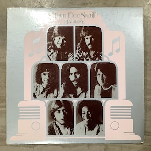 May include: Album cover for Three Dog Night's "Harmony." The album art features a light blue background with a pink radio-like design. Six band member portraits are arranged in the center. The band's name and album title are at the top.