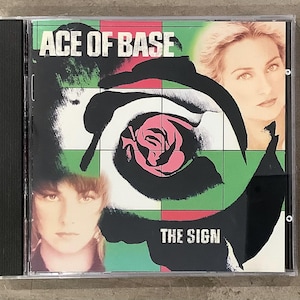 May include: A CD case for the album "The Sign" by Ace of Base. The cover features a stylized rose with black and white swirls, pink petals, and the band's name in white. Two women's faces are visible.