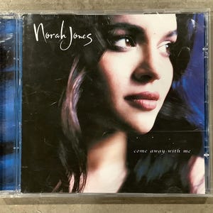 May include: CD case featuring Norah Jones with the album title "come away with me." The cover art showcases a close-up of Jones' face with dark hair and a blue background. The artist's name is written in cursive.