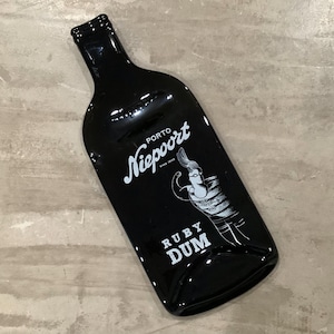 May include: A black glass bottle with white text and a graphic of a woman. The bottle reads "Niepoort PORTO" and "RUBY DUM". The bottle is on a light gray surface.