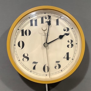 May include: A round wall clock with a mustard yellow frame and an off-white face. The clock features large black serif numerals from one to twelve, black hour and minute hands, a thin silver second hand, and the Sunbeam brand name printed in black.