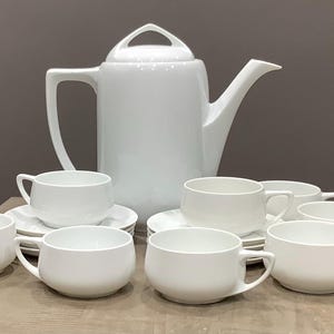 May include: A white porcelain tea set, comprising a teapot and several teacups with saucers. The teapot has a handle and a spout. The cups are round with handles. The set is arranged on a light-coloured surface.