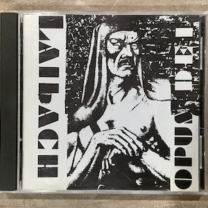 May include: A CD case featuring a black and white image of a person with a stern expression. The text "LAIBACH" is vertically aligned on the left, and "OPUS DEI" is on the right. The CD case is in a clear plastic sleeve.
