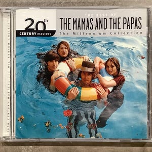 Peut inclure: Un boîtier de CD avec l'album "The Best of The Mamas and The Papas". La pochette montre les membres du groupe dans une piscine avec une bouée gonflable colorée. Le texte "20th Century Masters" est visible.
