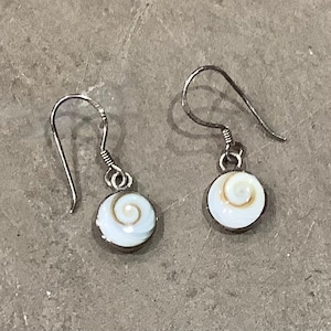 May include: A pair of drop earrings featuring circular white shell charms with a natural brown spiral pattern. Each charm is set in a simple metal frame and hangs from a curved silver-toned ear wire, creating a delicate and balanced jewelry piece.