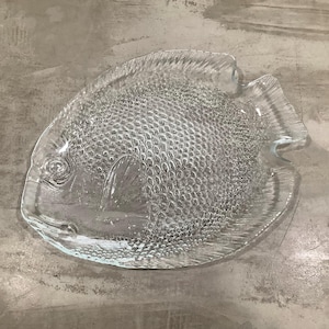May include: A clear glass fish-shaped serving dish with a textured surface. The dish is designed to resemble a fish with scales and a raised fin.