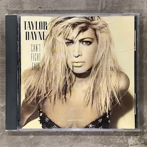 Może przedstawiać: Pudełko na płytę CD z albumem Taylor Dayne "Can't Fight Fate". Okładka albumu przedstawia portret kobiety z blond włosami. Tekst "TAYLOR DAYNE" i "CAN'T FIGHT FATE" jest wydrukowany na czarno w lewym górnym rogu.