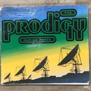May include: Vintage album cover for "The Prodigy - Out of Space." The design features the band's name in bold, black letters with a green outline, set against a gradient sky. Silhouetted satellite dishes are in the foreground.