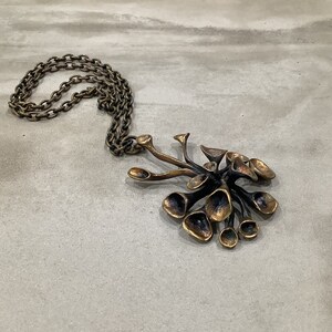 May include: A gold-toned necklace with a large, abstract pendant. The pendant is made of multiple, interconnected, organic shapes that resemble a flower or a cluster of leaves.
