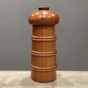 Może przedstawiać: Wysoki drewniany młynek do pieprzu z zaokrągloną górą i cylindrycznym korpusem. Młynek wykonany jest z jasnobrązowego drewna i ma gładkie, wypolerowane wykończenie. Korpus podzielony jest na sekcje poziomymi pasmami.