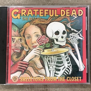 May include: A CD case featuring the album "The Best of Grateful Dead: Skeletons from the Closet." The cover art includes a woman holding a rose, a skeleton, and the band's name in orange lettering. The background is a mix of red, blue, and green.