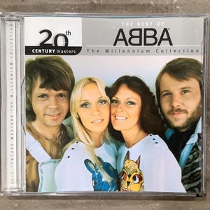 May include: A CD case for The Best of ABBA The Millennium Collection, 20th Century Masters. The cover features a photograph of the four band members. The two women in the centre wear white shirts with colourful graphic prints, while the men wear casual tops.