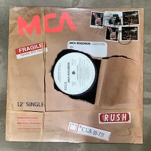 May include: A vintage 12" single record in a brown cardboard mailer. The mailer has red "MCA" lettering, a "Fragile" sticker, and a "Rush" label. The record label is visible through a torn opening, with the text "MCA Records".