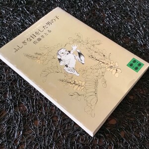 May include: A beige book with a black and white illustration of a creature with a green leaf on the cover. The book title is "ふしぎな日々を して た の は" and the subtitle is "花 織 さ と"