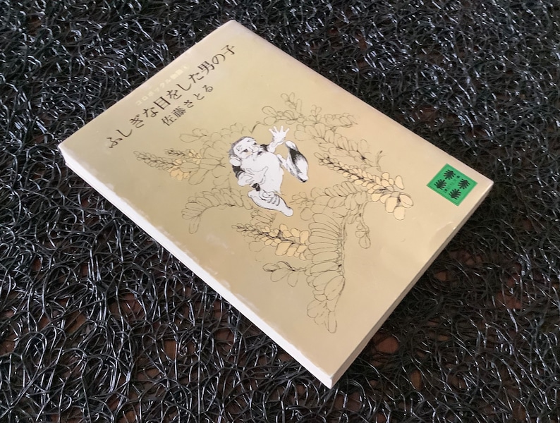 May include: A beige book with a black and white illustration of a creature with a green leaf on the cover. The book title is "ふしぎな日々を して た の は" and the subtitle is "花 織 さ と"