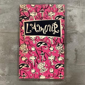 May include: A vintage book cover with a pink background and a repeating pattern of black and white abstract designs, including masks, flowers, and champagne glasses. The title "L'AMOUR" is displayed in a black rectangle.