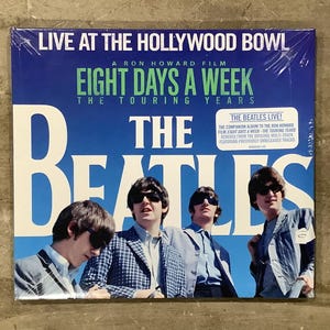 May include: A vinyl record cover for The Beatles Live at the Hollywood Bowl, featuring a blue background with white text and a photograph of the four band members wearing sunglasses and jackets. The cover includes the text Eight Days a Week.