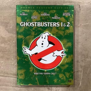 May include: A Ghostbusters 1 & 2 double feature DVD gift set with a green background featuring a white ghost character inside a red circle with a diagonal line. The text reads Bill Murray, Dan Aykroyd, Sigourney Weaver, Harold Ramis, Rick Moranis, and Who You Gonna Call?