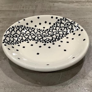May include: A small, white ceramic dish with a decorative black pattern. The pattern consists of a series of interconnected shapes and scattered dots. The dish is oval-shaped and has a slightly raised edge.