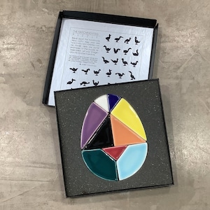 May include: A colorful, handmade egg puzzle with geometric shapes in a black box. The puzzle pieces are various colors, including blue, yellow, purple, and red. The box lid has text that reads "THE HATCHING EGG PUZZLE".