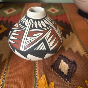 May include: A small, round, hand-painted ceramic vase with a white rim. The vase features a geometric design in black, white, and reddish-brown. The vase is sitting on a colorful woven textile.