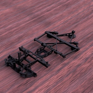 Op de afbeelding: Twee zwarte componenten van een modeltrein chassis worden getoond op een roodbruin houten oppervlak. De componenten hebben ingewikkelde geometrische ontwerpen met rechthoekige en schuine steunen en kleine wielen.
