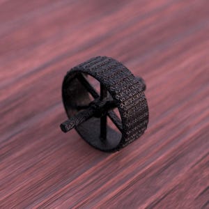 Op de afbeelding: Een zwart, getextureerd wiel met spaken en een centrale as. Het wiel heeft een dikke, bandachtige buitenband. De achtergrond is een vage, roodbruine ondergrond.