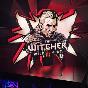 Può includere: Un'insegna decorativa con il Witcher della serie di videogiochi. L'insegna ha un design a stella rossa e bianca con la scritta "The Witcher Wild Hunt". L'immagine è ambientata su uno sfondo scuro.