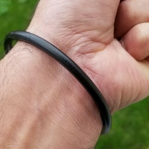 May include: A black, circular bracelet made of a smooth material. The bracelet is worn on a wrist, and the image is a close-up shot. The bracelet appears to be a simple, minimalist design.