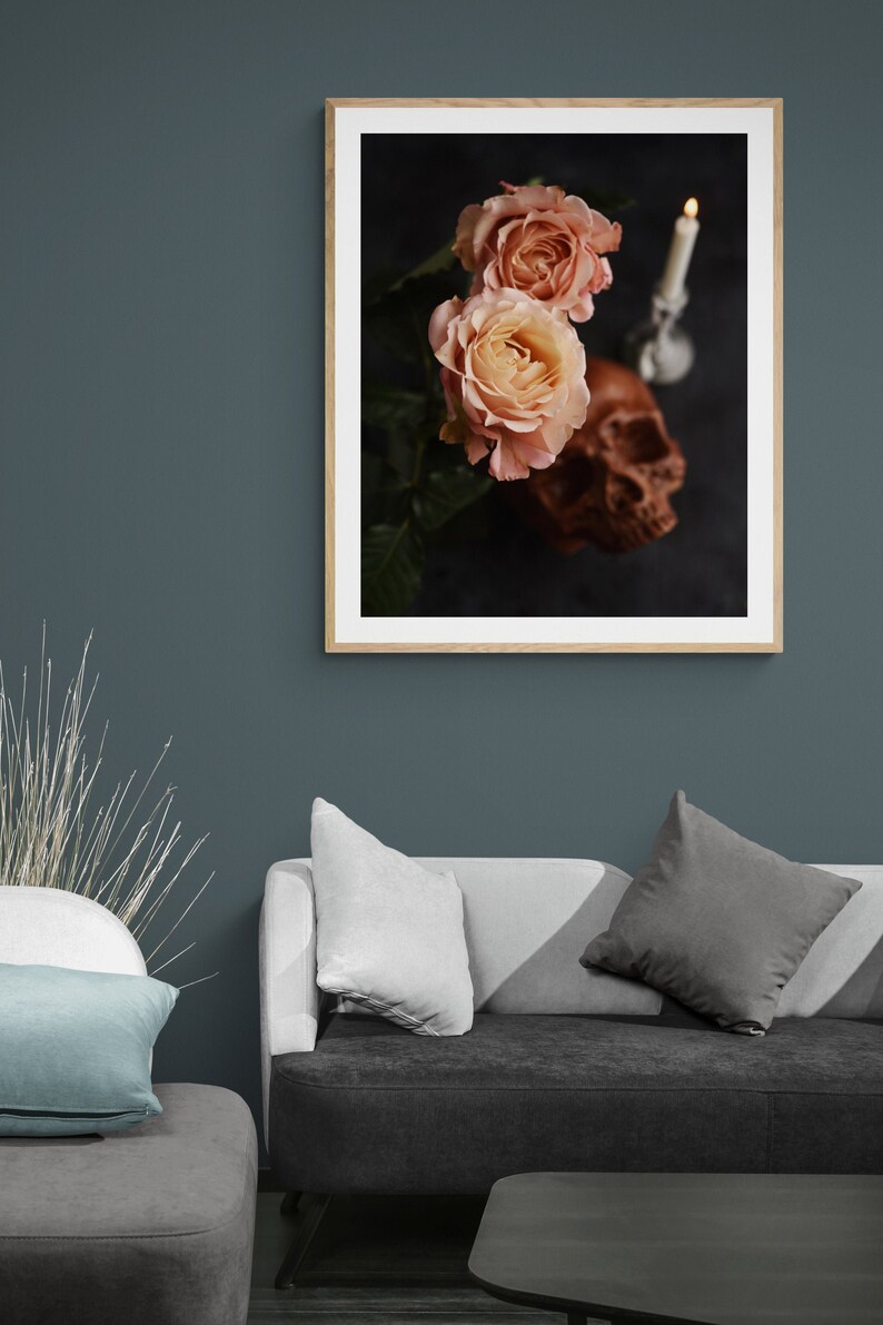 May include: A framed print showing two peach roses, a brown skull, and a lit candle against a dark backdrop. The artwork has a gothic or romantic theme. The frame is a light wood colour.