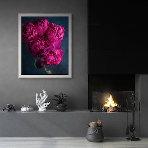 May include: A framed print of a bouquet of vibrant pink peonies against a dark blue background. The peonies are arranged in a vase with a gold handle. The print is hanging on a gray wall above a fireplace.