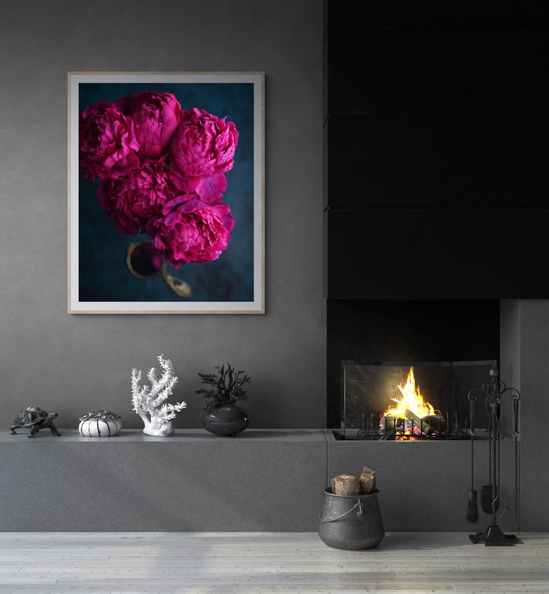 May include: A framed print of a bouquet of vibrant pink peonies against a dark blue background. The peonies are arranged in a vase with a gold handle. The print is hanging on a gray wall above a fireplace.