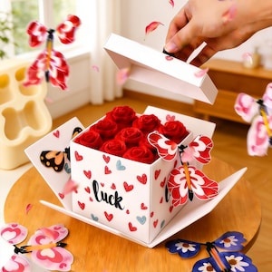 Może przedstawiać: Białe pudełko na prezent ozdobione czerwonymi i niebieskimi sercami oraz napisem "luck" zawiera czerwone róże. Pudełko otoczone jest dekoracyjnymi zabawkami w kształcie motyli z czerwono-białymi skrzydłami. Ręka otwiera pudełko.