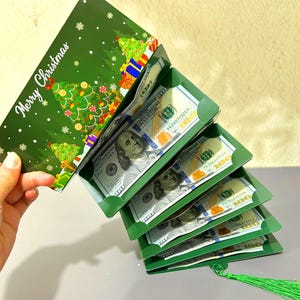 Puede incluir: Un tarjetero verde con temática navideña con las palabras "Merry Christmas" y un diseño de árbol de Navidad. El tarjetero contiene varios billetes de 100 dólares, visibles en los compartimentos abiertos. Una borla verde cuelga de la parte inferior.