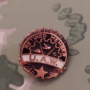 May include: A copper-colored pin with a black and white enamel design. The pin features a gear with a plane and a car inside, a white banner with the text "U.A.W." and the words "Retired Member" around the outside. The pin has a star at the bottom.