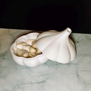 Könnte beinhalten: Ein weißer, knoblauchförmiger Keramikbehälter mit Deckel, der teilweise geöffnet ist, um Knoblauchzehen im Inneren freizulegen. Der Behälter steht auf einer hellgrünen und weißen Oberfläche mit schwarzem Hintergrund.