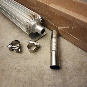 May include: An assortment of metallic automotive parts, including a ribbed flexible pipe, two clamps, and a finned heat sink. The components are silver and arranged on a concrete surface near a cardboard box.