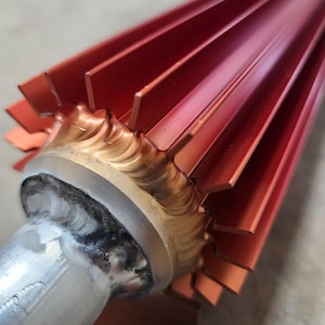 May include: Close-up of a metallic red industrial component. The component features a series of parallel fins, with a welded gold-colored base and a silver-colored cylindrical shaft. The fins are evenly spaced and angled.