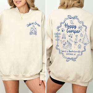 K&ouml;nnte beinhalten: Cremefarbenes Sweatshirt mit blauen Grafiken. Auf der Vorderseite steht "Cabin Crew" &uuml;ber einer Lagerfeuer-Illustration. Auf der R&uuml;ckseite steht "Happy Camper" in einem dekorativen Rahmen, umgeben von Camping-Symbolen und Text: "Carol's Bachelorette Scottsdale, AZ".