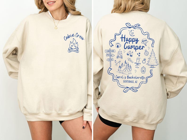 K&ouml;nnte beinhalten: Cremefarbenes Sweatshirt mit blauen Grafiken. Auf der Vorderseite steht "Cabin Crew" &uuml;ber einer Lagerfeuer-Illustration. Auf der R&uuml;ckseite steht "Happy Camper" in einem dekorativen Rahmen, umgeben von Camping-Symbolen und Text: "Carol's Bachelorette Scottsdale, AZ".